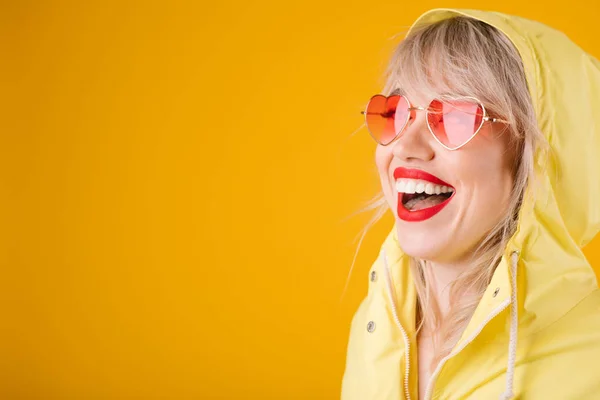 Impermeable amarillo. Feliz mujer riendo sobre fondo amarillo witn gafas de sol en forma de corazón rosa. Emociones brillantes —  Fotos de Stock