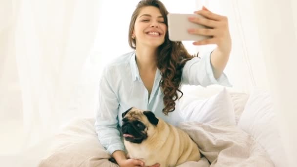 Mujer en la cama tomando selfie con su pequeño perro pug. Imágenes de vídeo — Vídeo de stock