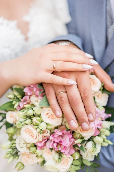 Handen van de bruid en bruidegom close-up, het dragen van witgouden trouwringen om haar handen, de bruid houdt een bruiloft boeket van roze rozen met veren, linten en versierd met steen — Stockfoto