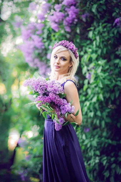 一个被紫色花朵环绕的年轻漂亮女子的户外时尚照片 穿着长裙的女孩们 背景上有一个狭缝 后面是一个春天的花园 有紫丁香 化妆品和香水的概念 你头发上的丁香花束和花朵 — 图库照片
