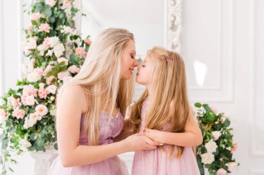 Zarif elbiseli, güzel ve mutlu bir sarışın anne, bahar sezonunun arka planında çiçeklerle küçük kızına sarılıyor. Güzel parlak stüdyo çiçeklerle süslenmiş. Şenlik havası