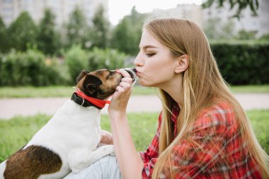 young pretty girl in red tartan shirt and jeans sit at the lawn near high buildings and kiss her dog. Love and friendship concept. Profile view, close up portrait.