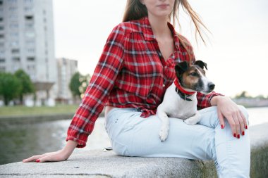 Young woman seat on the embankment near high-rise building. City background. Small fox terrier lies on her lap.