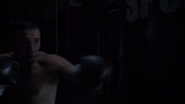 La cámara se mueve alrededor de un saco de boxeo y un boxeador asertivamente golpeando en un viejo gimnasio — Vídeo de stock
