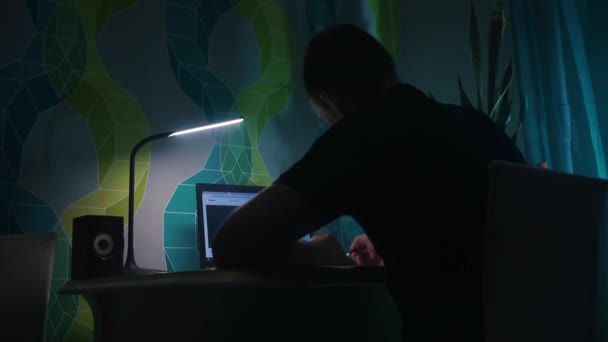 Guy sits table and writes, in front laptop. Evening, night, apartment table lamp — Stock Video