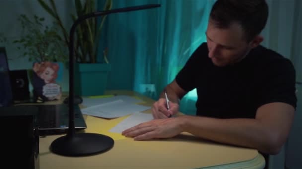 Парень думает, что написать, в письме на бумаге, сидя за столом, с ручкой в руках — стоковое видео
