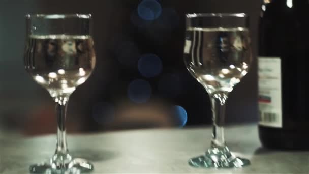 Camera moves back and forth between 2 glasses, bottle of champagne on table. Flashing lights, christmas tree on background — Stock Video