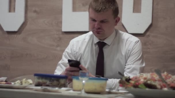 Cara em um feriado sentado em uma mesa com comida, olhando para um smartphone — Vídeo de Stock