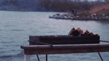 In nature, logs burn in the brazier, smoke against the background of the river, shore.