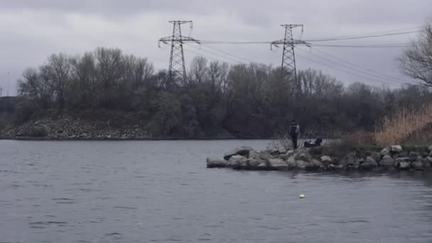 Cloudy, dull day, man jacket stands on the banks river, power lines background — Stock video