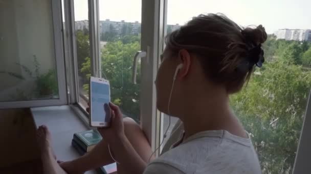 Het meisje met de koptelefoon leunde met haar hoofd tegen het raam op balkon, vensterbank — Stockvideo