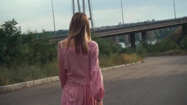 ピンクのドレスを着た女の子が橋の下の放棄された道を一人で歩いている — ストック動画
