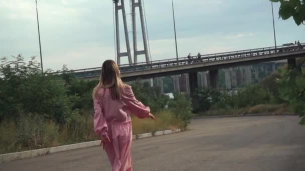 Dívka v růžových šatech, kráčí sama po opuštěné cestě pod mostem — Stock video