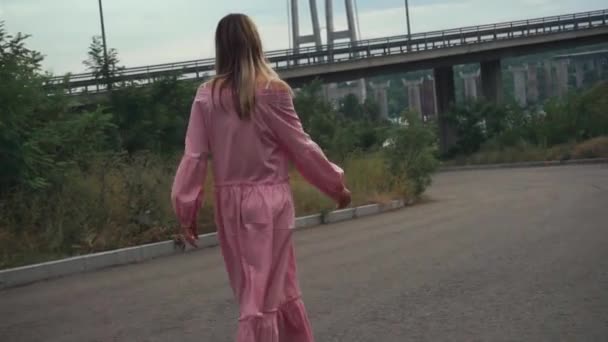 Een meisje in een roze jurk, loopt alleen over een verlaten weg onder een brug — Stockvideo