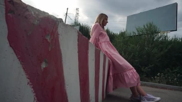Дівчина в рожевій сукні сидить на бетонному блоці, порожній рекламний щит на фоні — стокове відео