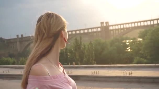 Ein fröhliches, fröhliches, lächelndes Mädchen in einem Kleid schreitet auf der Brücke, der Sonne gegenüber — Stockvideo