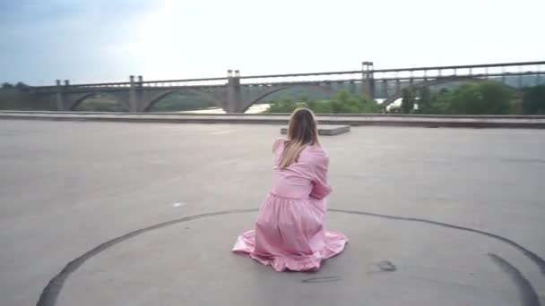 Menina em um vestido de cócoras perto de uma ponte rodoviária à noite olha para o pôr do sol — Vídeo de Stock