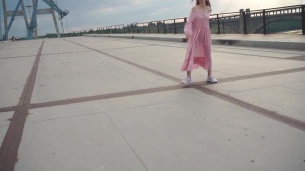 Радісна дівчина в сукні, що крутиться навколо своєї осі на бетонному залізному мосту — стокове відео