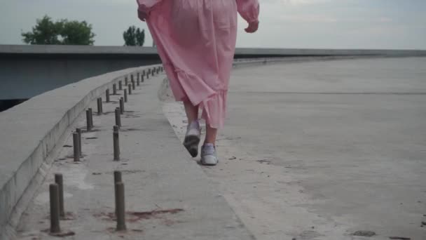 Het meisje alleen loopt over een verlaten, betonnen weg, een onvoltooide brug — Stockvideo