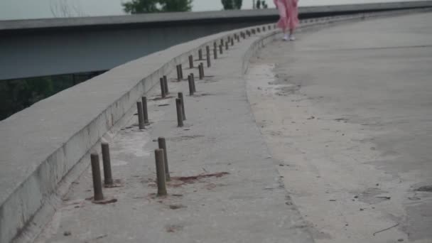 A lány egyedül sétál egy elhagyatott, betonozott úton, egy befejezetlen hídon. — Stock videók
