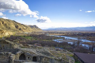 Uplistsikhe mağaraları, Georgia. Kasım 2017 civarı. Bir turistin dağları, nehir ve taş devri mağaraları olan muhteşem mavi gökyüzü altında bulutlu güzel bir köy manzarası..