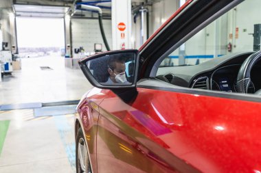 Beyaz tenli genç bir teknisyen, virüsün yayılmasını önlemek için maske ve eldivenle korunan bir aracın içinde güvenlik incelemesi yapıyor.