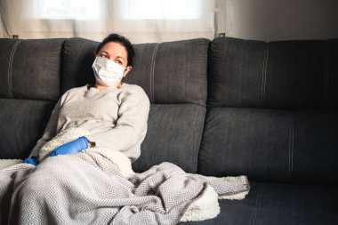 Maskeli ve tıbbi eldivenli hasta kadın kanepeye oturmuş battaniyeyle kaplanmış, muhtemelen kovid-19 'dan etkilenmiş.