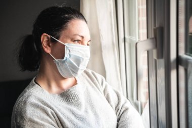 Maskeli ve tıbbi eldivenli hasta kadın pencereden bakıyor. Muhtemelen koronavirüs enfeksiyonu kapmış ve kendini izole etmiş.