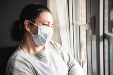 Maskeli ve tıbbi eldivenli hasta kadın pencereden bakıyor. Muhtemelen koronavirüs enfeksiyonu kapmış ve kendini izole etmiş.
