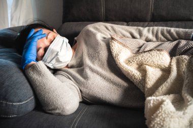 Kanepede yatan yüzü maskeli ve tıbbi eldivenli hasta kadın. Muhtemelen koronavirüs enfeksiyonu kapmış.