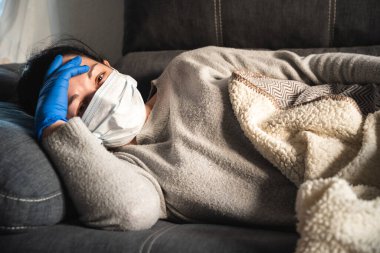 Kanepede yatan yüzü maskeli ve tıbbi eldivenli hasta kadın. Muhtemelen koronavirüs enfeksiyonu kapmış.