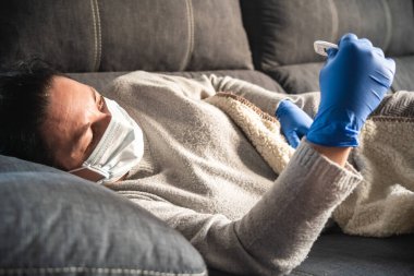 Yüz maskesi ve tıbbi eldivenleri olan hasta bir kadın kanepede uzanmış termometreye bakıyor, muhtemelen koronavirüsten etkilenmiş.
