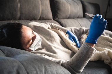 Yüz maskesi ve tıbbi eldivenleri olan hasta bir kadın kanepede uzanmış termometreye bakıyor, muhtemelen koronavirüsten etkilenmiş.