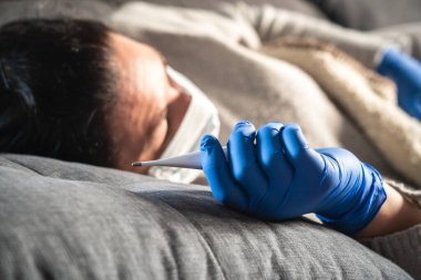 Kanepede uyurken elinde termometre tutan maskeli ve tıbbi eldivenli hasta bir kadının eli. Muhtemelen koronavirüsten etkilenmiştir.