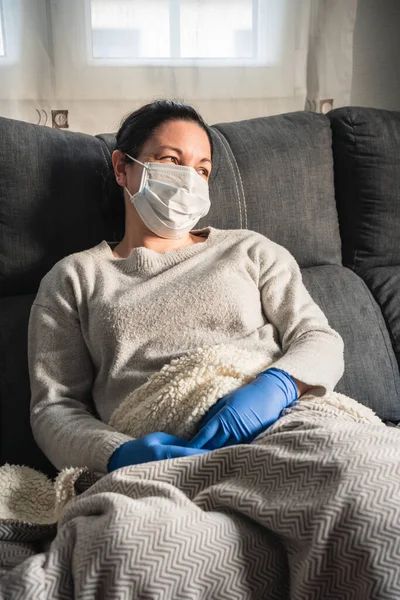 Maskeli ve tıbbi eldivenli hasta kadın kanepeye oturmuş battaniyeyle kaplanmış, muhtemelen kovid-19 'dan etkilenmiş.