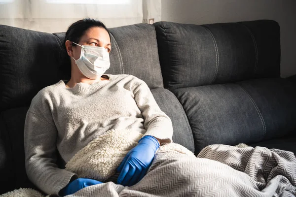 Sick woman with mask and medical gloves sitting on sofa covered with blanket, probably infected with covid-19
