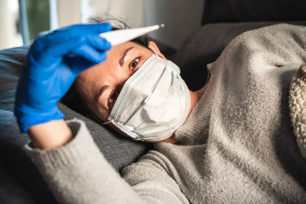 Sick woman with face mask and medical gloves looking at a thermometer lying on the sofa, probably infected by coronavirus