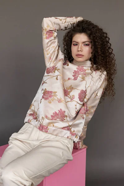 Young beautiful Caucasian with afro curls hairstyle wearing casual outfit  seating on pink box on grey background