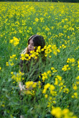 Sarı tecavüz tarlasında oturan koyu renk elbiseli, güzel saçlı bir kız. Ilık bahar günü