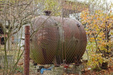 Bitki örtüsündeki bir çitin arkasında kahverengi paslı bir demir tank.
