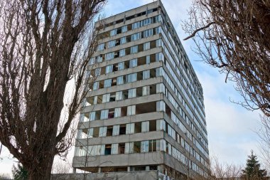 Kuru ağaçların ve mavi gökyüzünün arka planında kırık camları olan büyük, gri terk edilmiş bir ev.