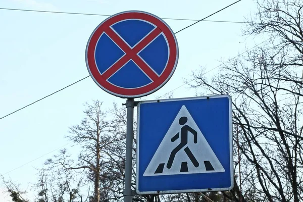 two road signs from a stop is prohibited and a pedestrian crossing on an iron pole against the background of branches and a blue sky on a city street