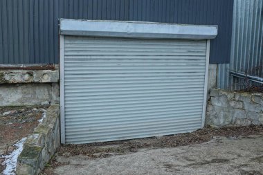 Eski bir garajın ön cephesinde beyaz plastik kapılar ve gri bir yolun kenarında kepenkleri var.