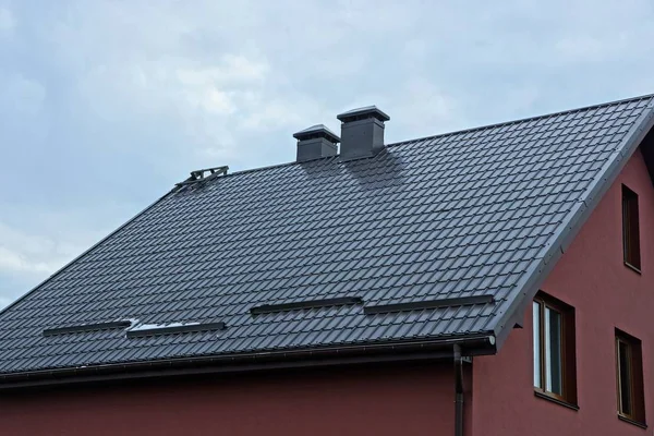 part of a private house with a red wall under a brown tiled roof with metal chimneys against a background of gray sky and clouds