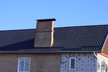 Evin bir kısmı, kahverengi kiremitli çatının altında bir duvar ve pencereyle büyük tuğladan bir baca, mavi gökyüzüne karşı.