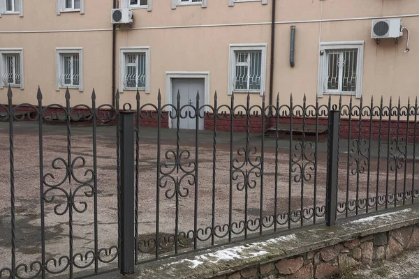 black metal fence of sharp iron bars in a forged pattern on the street in front of a brown house with white windows