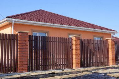 Güneşli bir günde mavi gökyüzüne karşı döşeli çatısı olan özel bir evin önündeki kahverengi tahta ve kırmızı tuğlalardan oluşan bir çit.