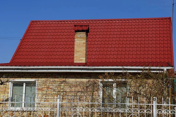 facade of a private house with a brown brick wall and windows under a red tiled roof and a long chimney against a blue sky on a sunny day