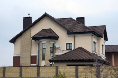 Çitin arkasındaki gri gökyüzüne karşı kiremitli çatı altında pencereleri olan büyük kahverengi bir ev. 