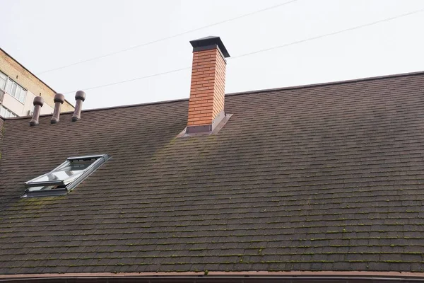 Kiremitli bir çatıdaki kahverengi tuğladan bir baca ve gri gökyüzünün arka planında bir pencere.